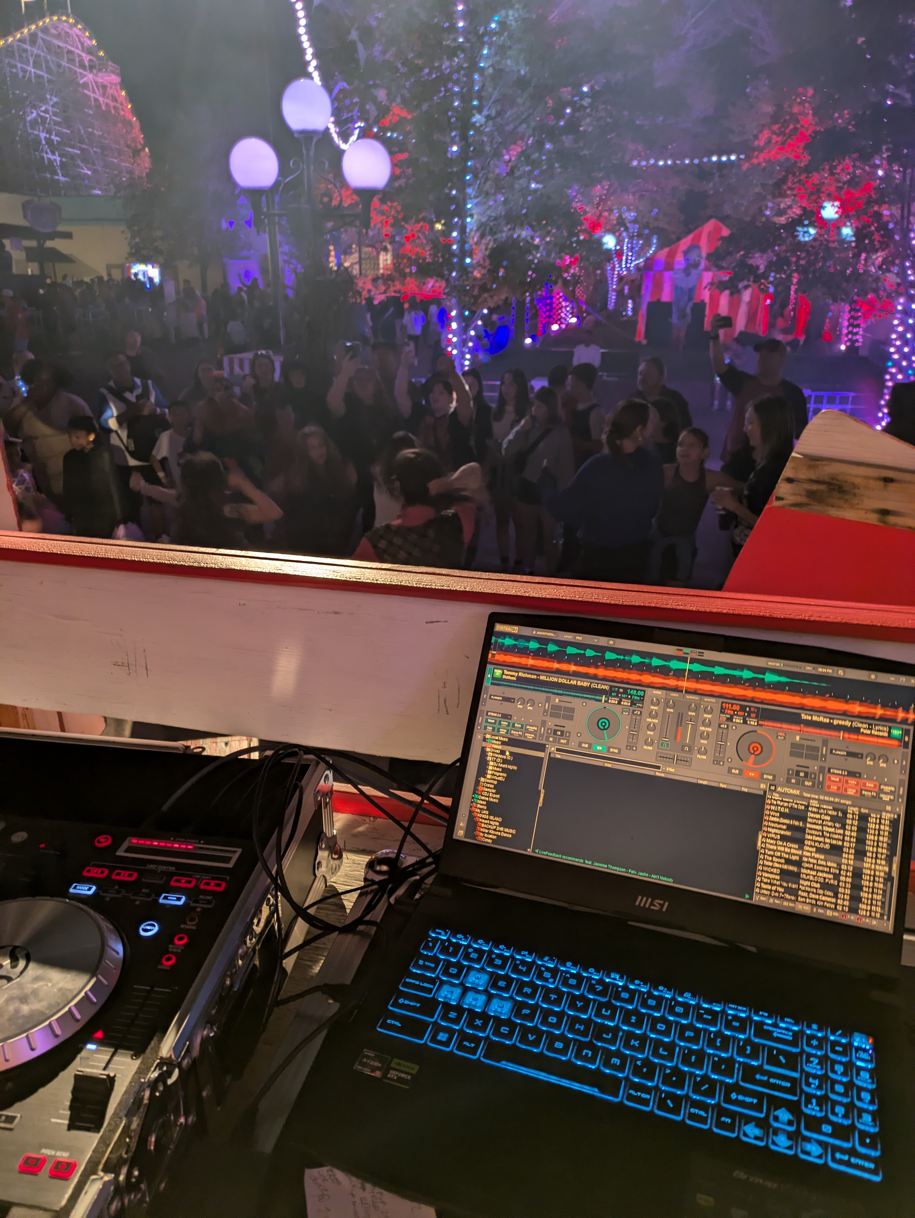 DJ booth with controller and laptop facing a packed night crowd under string lights